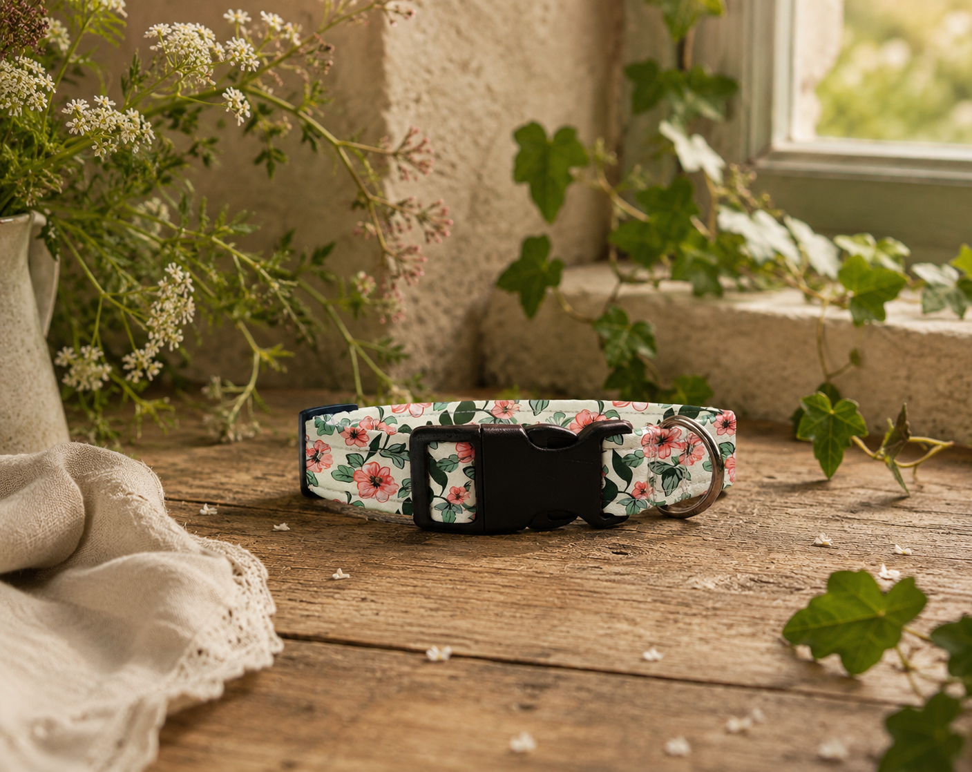 A floral dog collar with a black plastic buckle, resting on a rustic wooden surface surrounded by green ivy, small white flowers, and a beige cloth.